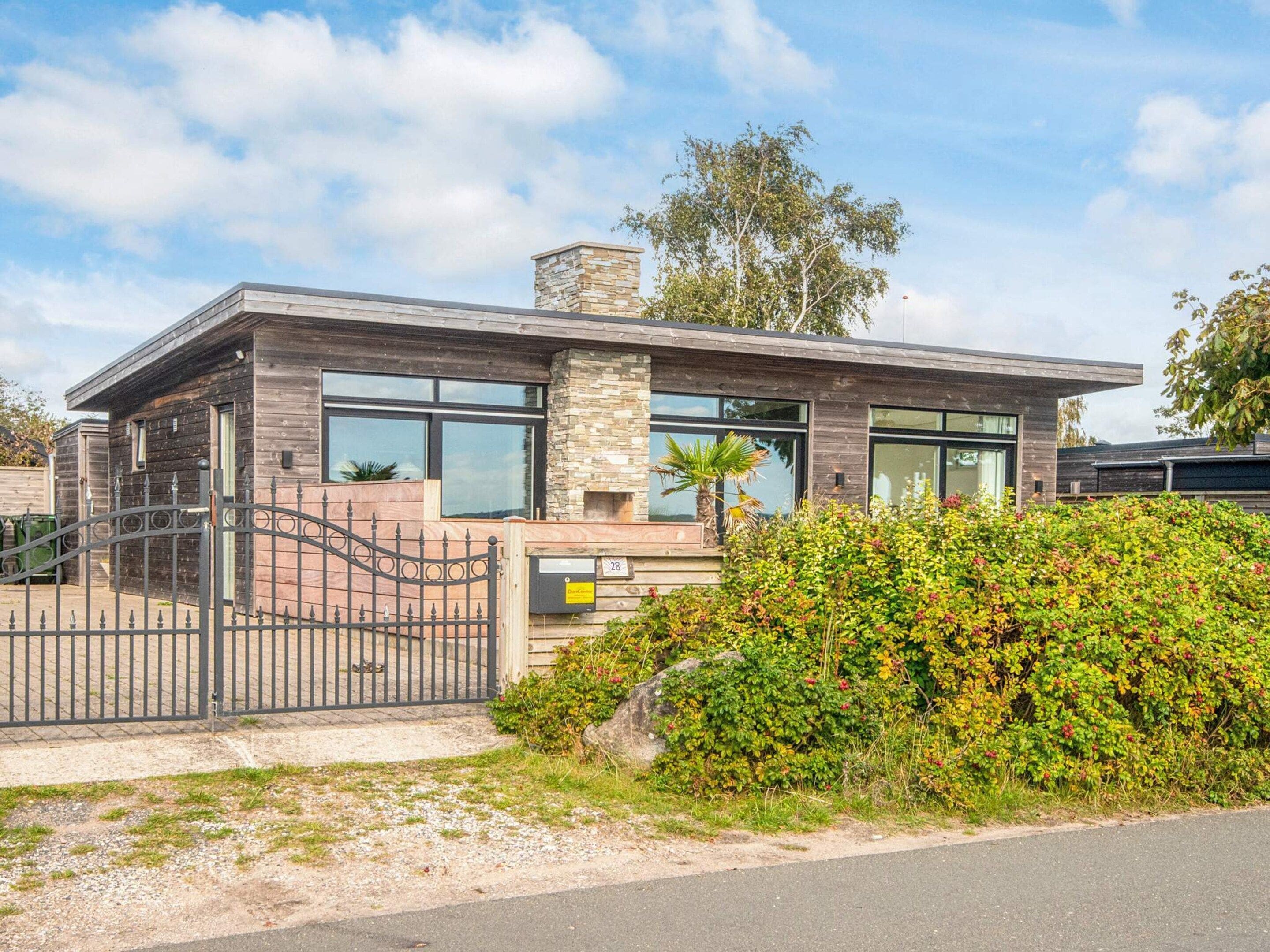 Une maison moderne à deux étages, dotée d’une cheminée en pierre, de grandes fenêtres et d’une entrée fermée par un portail.