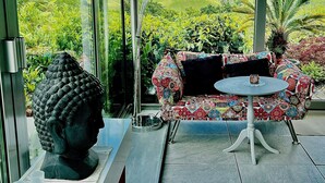 Et værelse med glasvæg, der giver udsigt over grønne marker og bakker. Indenfor er der en stribet sofa, et lille bord og en Buddha-figur.