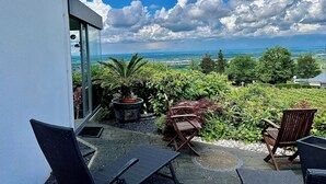 En balkon med udendørs møbler, en plantekasse og udsigt over et landskab med træer og en fjern horisont.
