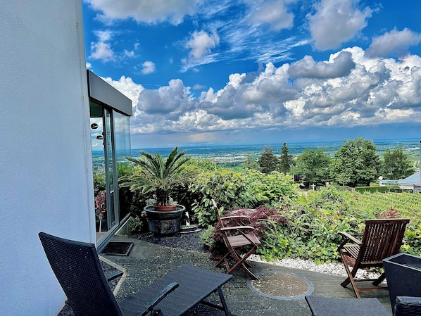 Ein Balkon mit Gartenmöbeln, einem Blumentopf und Blick auf eine Landschaft mit Bäumen und weiter Ferne.