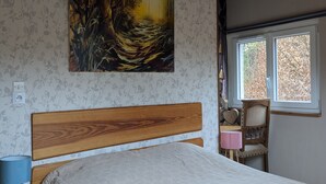 A bedroom with a wooden headboard, a painting of a forest scene, a window with a view of trees, and a bedside table with a lamp. - Equestrian Retreat in the Forest with Donkeys – Domaine L'échappée Verte (Soucht)