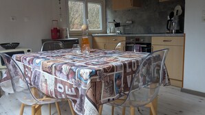 A dining area with a table covered by a quilt, surrounded by chairs, and a kitchen in the background. - Equestrian Retreat in the Forest with Donkeys – Domaine L'échappée Verte (Soucht)