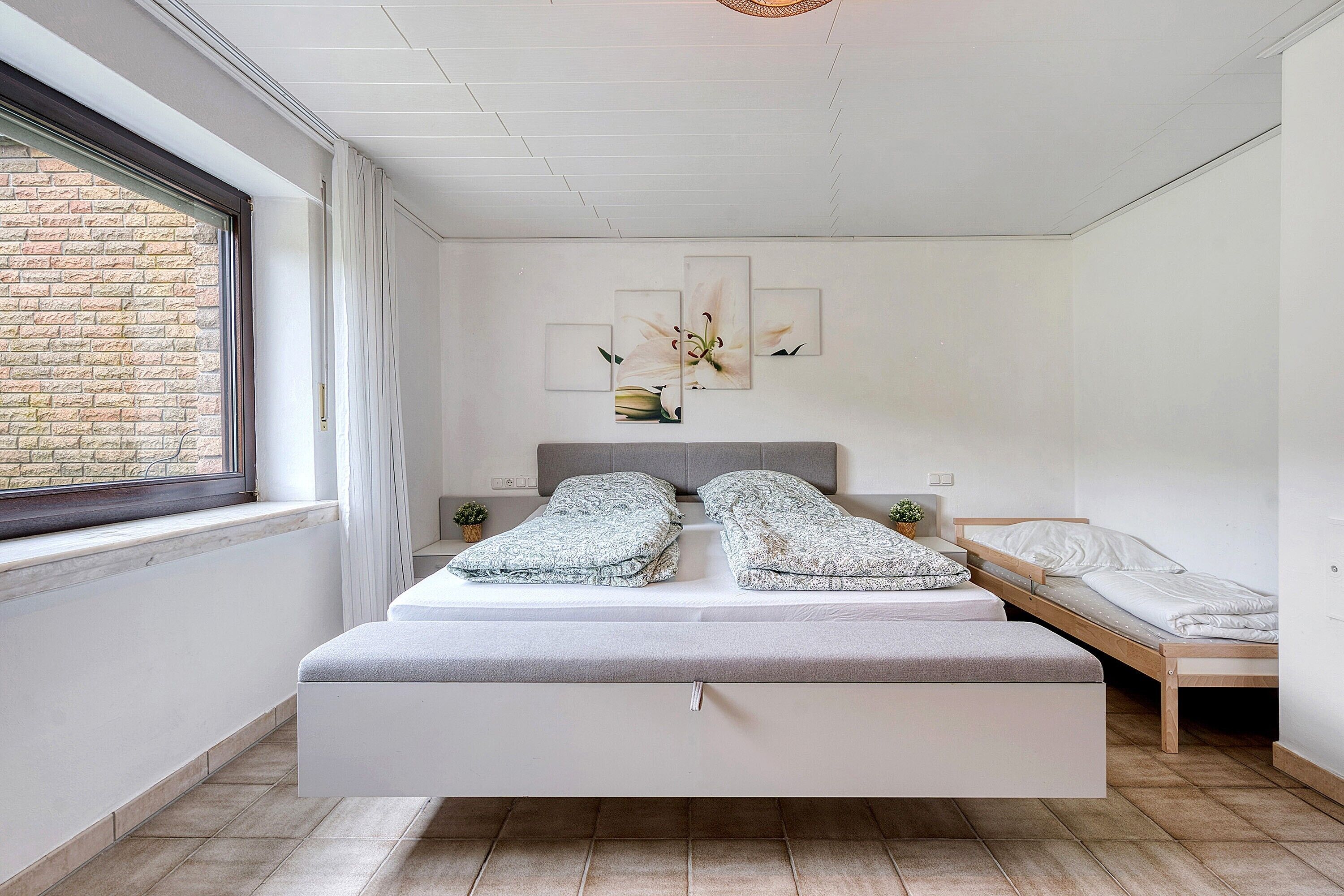A modern bedroom with a large bed, a smaller bed, a bench, a window, and a brick wall.
