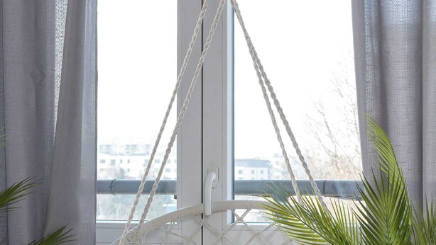 Ein weißer Makramee-Hängekorb mit Kissen, platziert neben einem Fenster mit Blick auf ein Gebäude und einen Zaun.