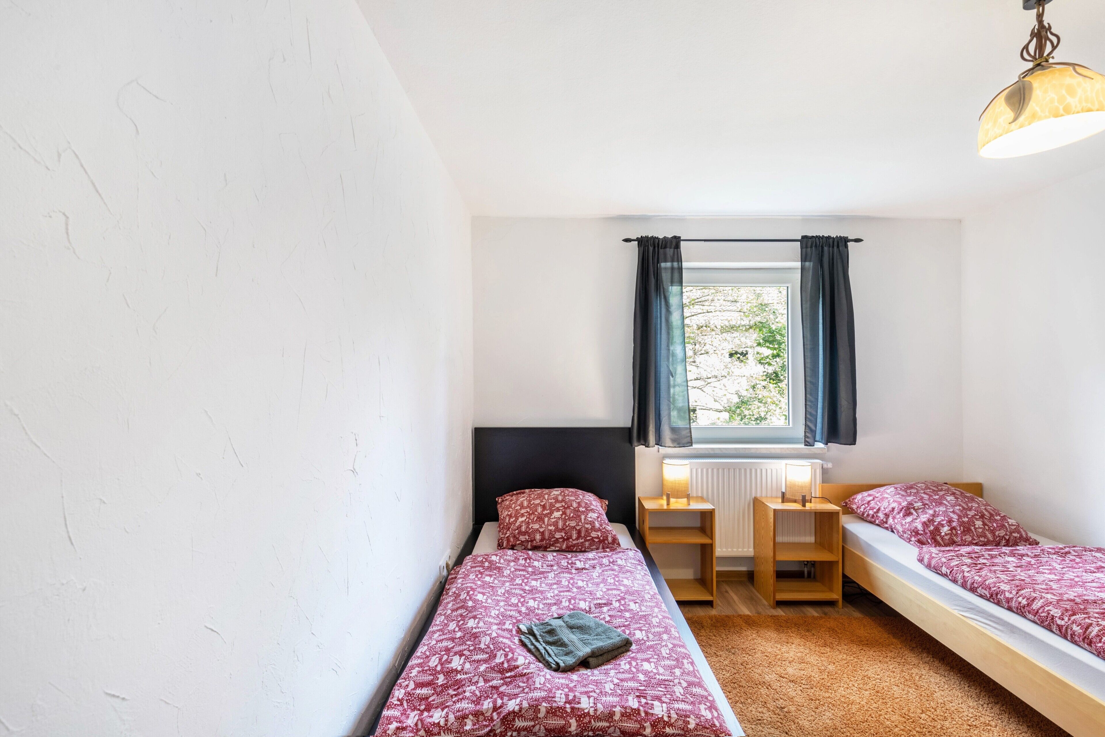 A small, single-bedroom room with a bed, a bedside table, a lamp, and a window with curtains.