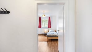 A narrow hallway leading to a bedroom with a bed, a window with curtains, a bedside lamp, and a radiator. - Apartment 'Yellow F' (Miltenberg)