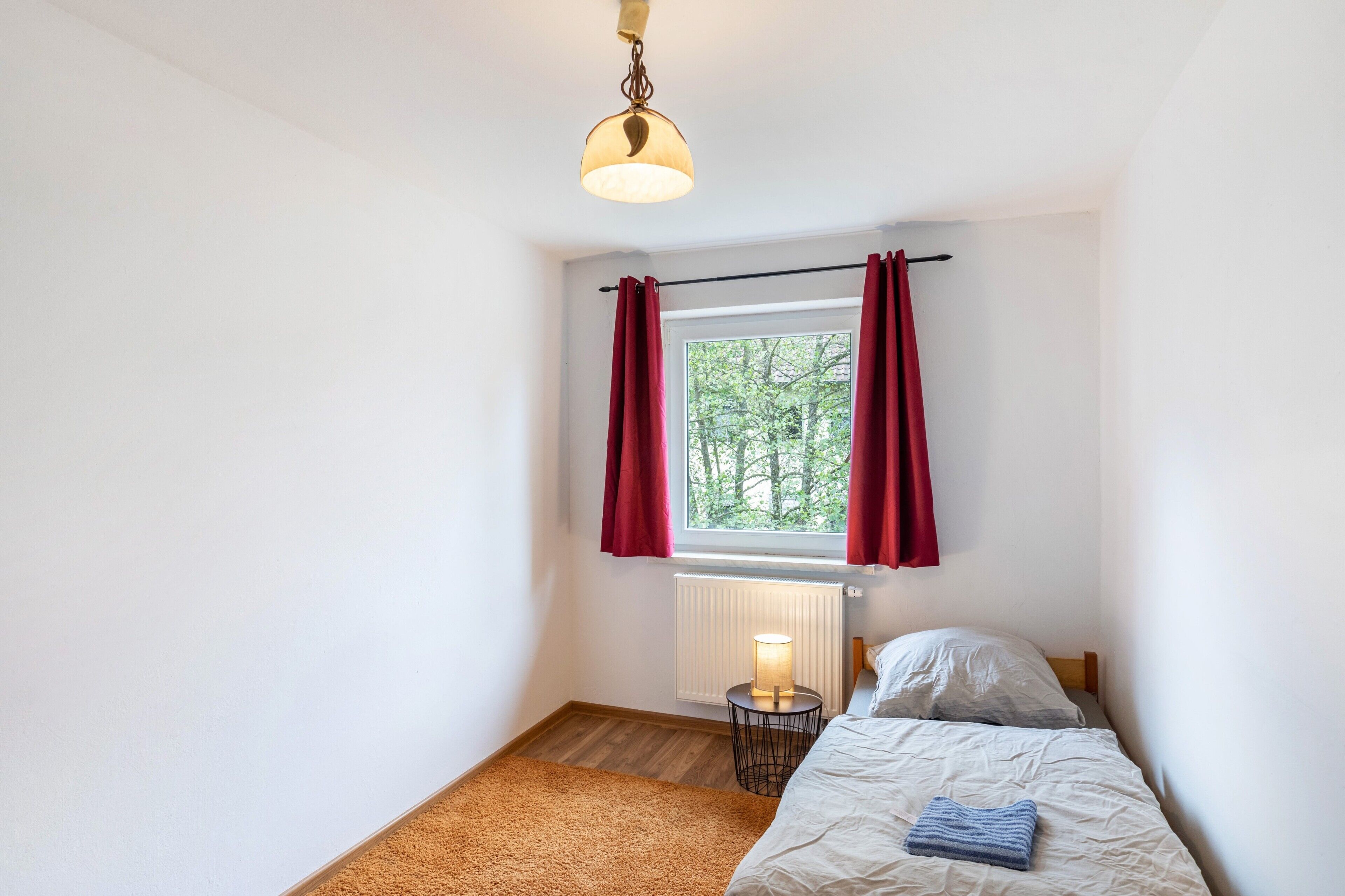 Une chambre à coucher avec un lit, une table de chevet, une lampe, une fenêtre avec des rideaux et un radiateur.