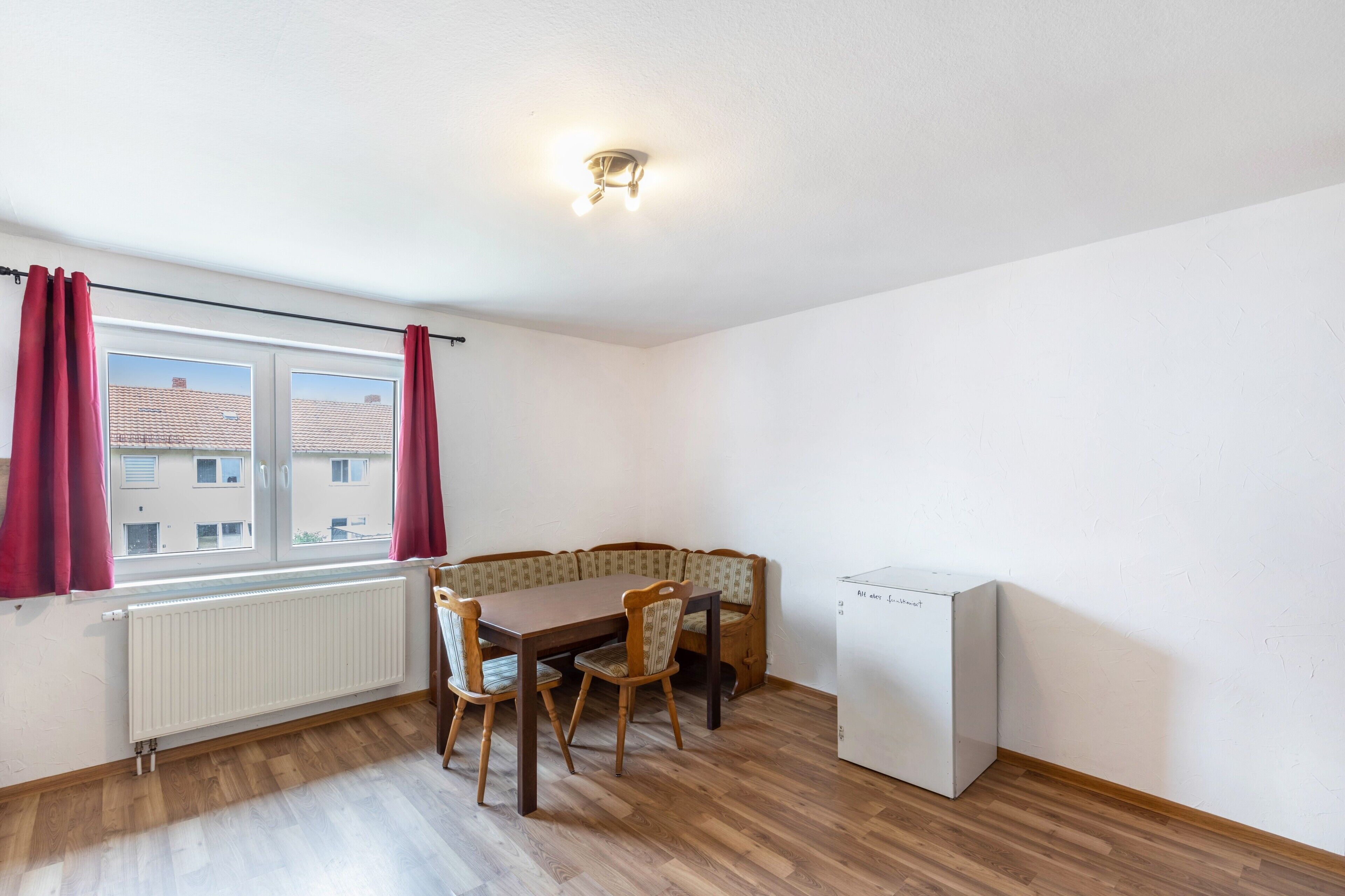 A room with a dining table, chairs, a bench, a radiator, a window with curtains, and a white appliance.