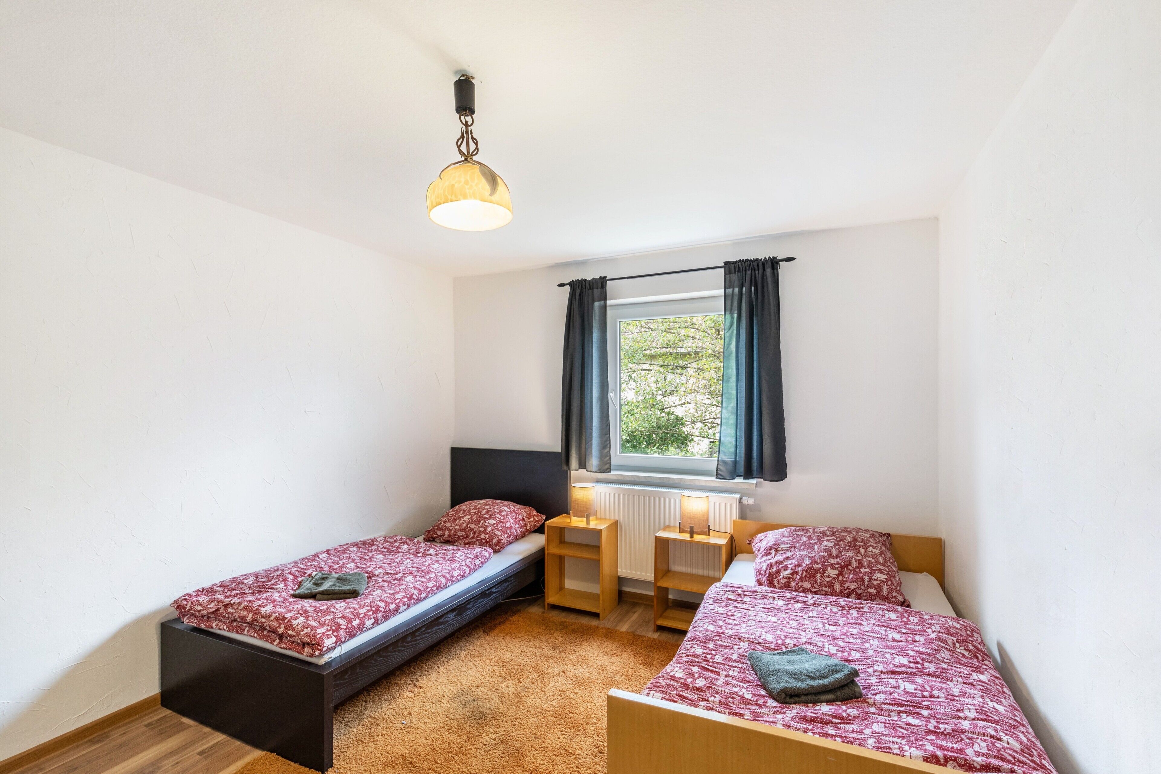 A small bedroom with two single beds, a window with curtains, a radiator, and a hanging light fixture.