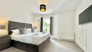 A bedroom with a large bed, a bedside table, a lamp, a radiator, and a TV mounted on the wall. - The Kipling Inn, 4 - Bed, Sleeps 6, Long Term Rental Available (Ulverston)