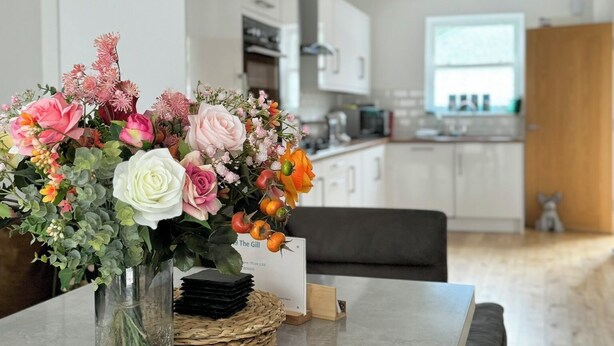 A modern kitchen with white cabinets, a dining table with a vase of flowers, and a remote control on the table. - The Kipling Inn, 4 - Bed, Sleeps 6, Long Term Rental Available (Ulverston)