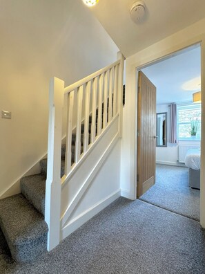 Eine Treppe mit weißen Geländern führt zu einem Schlafzimmer mit Holztür und einem Fenster mit Vorhängen.
