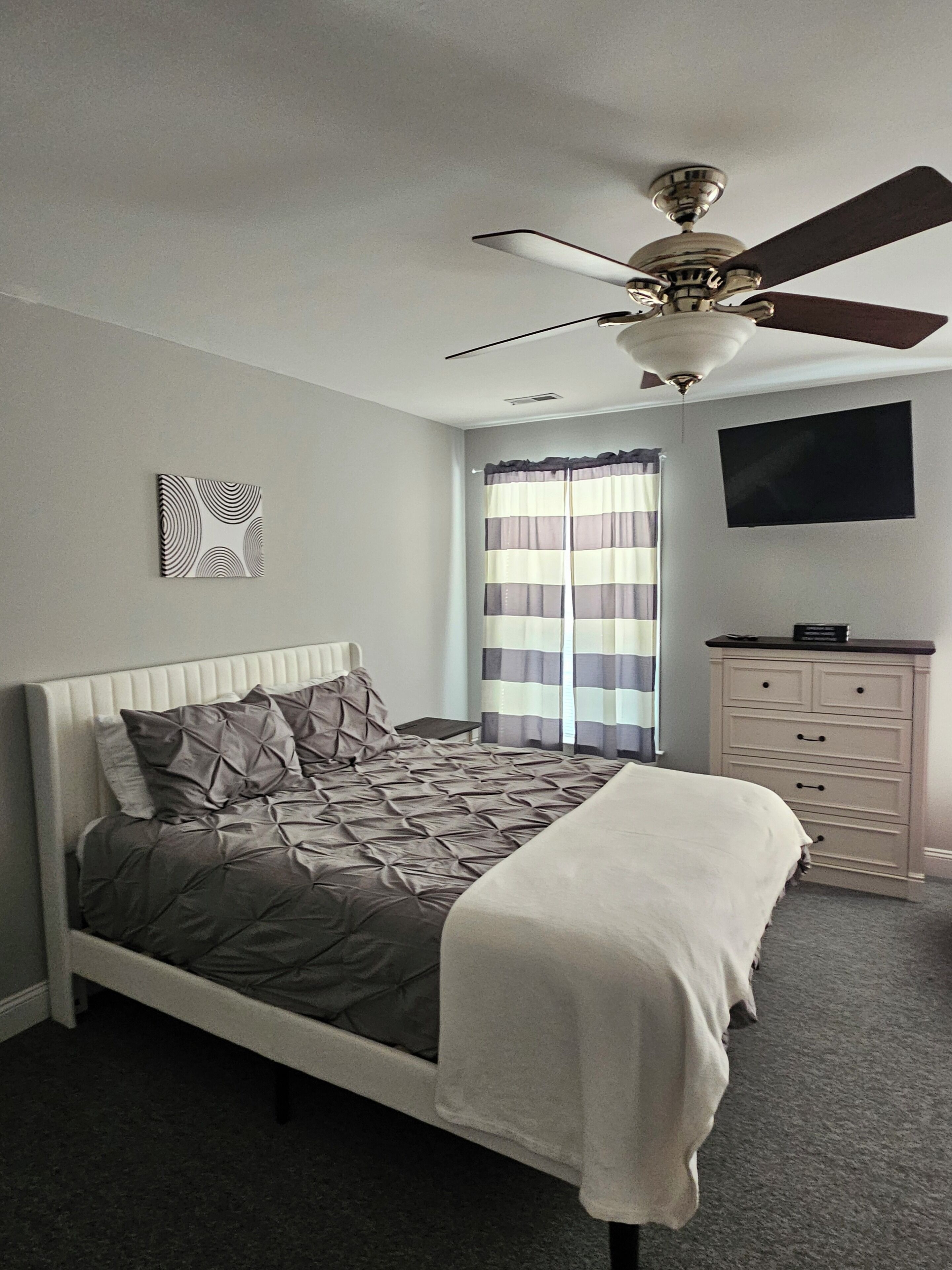 Une chambre à coucher avec un lit, un ventilateur de plafond, une télévision et une fenêtre avec des rideaux.