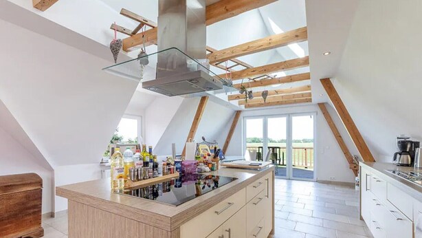 A modern kitchen with a central island, wooden beams, and a large sliding glass door to a balcony. - Holiday Home Kuhweid with Wellness Area, Private Terrace, Garden & Wi-Fi on Elbe Island Krautsand (Drochtersen)
