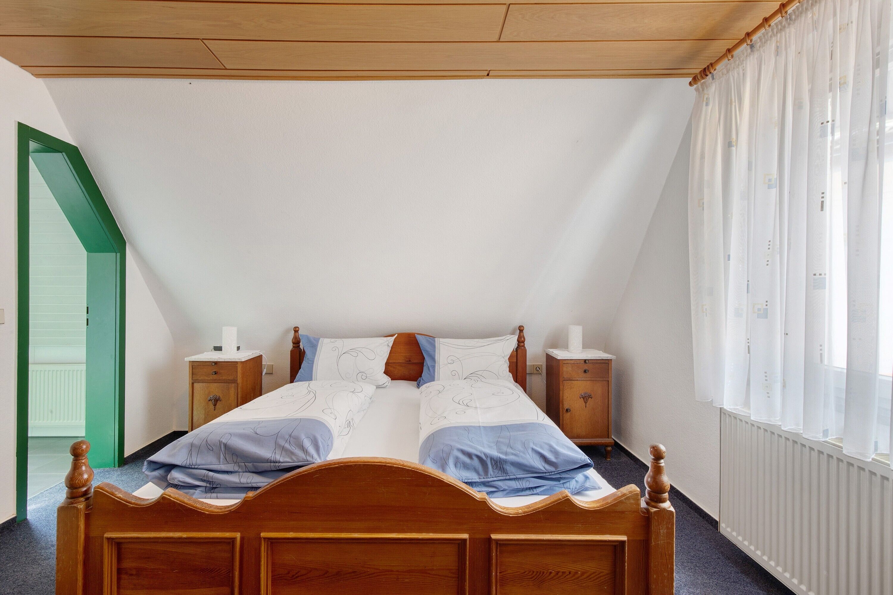 A bedroom with a wooden bed, two bedside tables, a radiator, and a window with curtains.