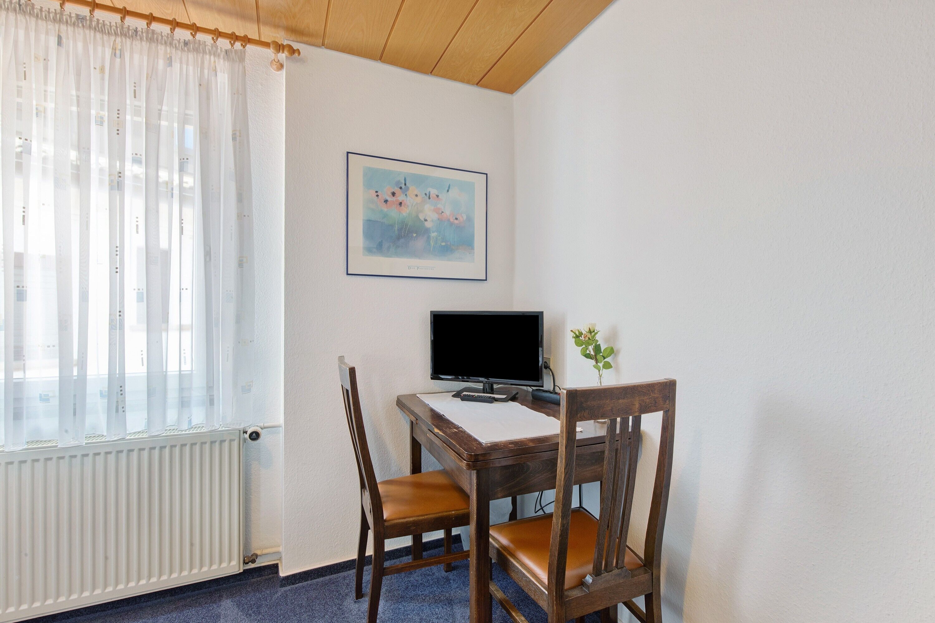 A room with a wooden desk, a computer monitor, two chairs, a radiator, a window with sheer curtains, and a framed picture on the wall.