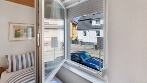 Een kamer met een bed, een raam met uitzicht op een straat en huizen, en een lijst met een afbeelding aan de muur.