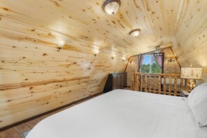 Et soveværelse i en træhytte med en seng, et vindue med gardiner og et natbord med en lampe.