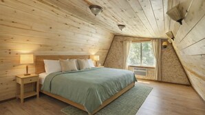 Ein gemütliches Schlafzimmer mit Holzbalkendecke, einem Bett mit grüner Tagesdecke, einem Nachttisch mit Lampe und einem Fenster mit Vorhängen.