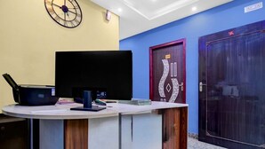 A reception desk with a flat-screen TV, a laptop, and some papers. There are two doors, one with a decorative design and another plain. A clock is on the wall. - Hotel O Metropolis Mall Kolkata (Barasat)
