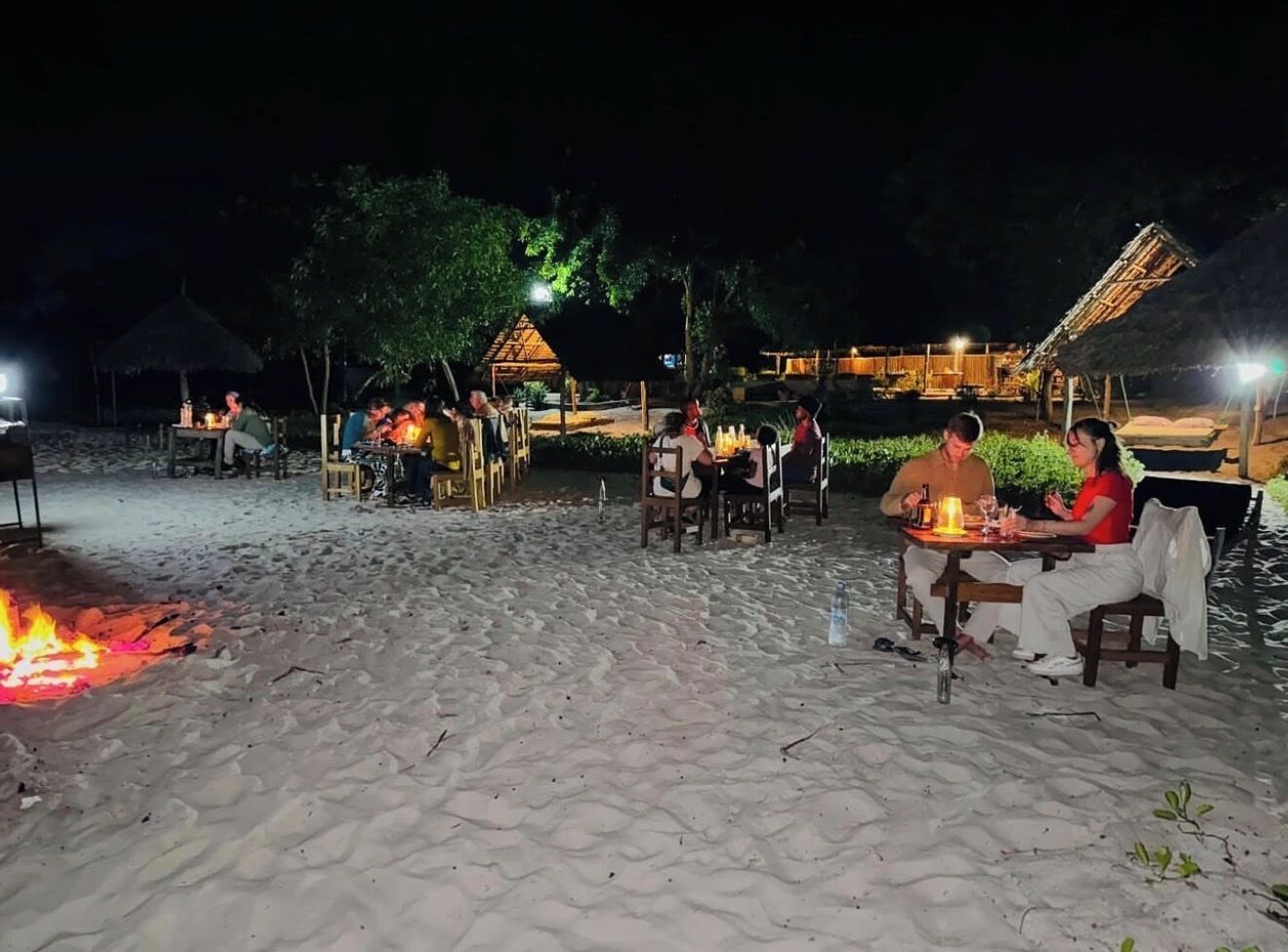 A beachside dining area with tables and chairs, a fire pit, and a thatched-roof structure in the background.