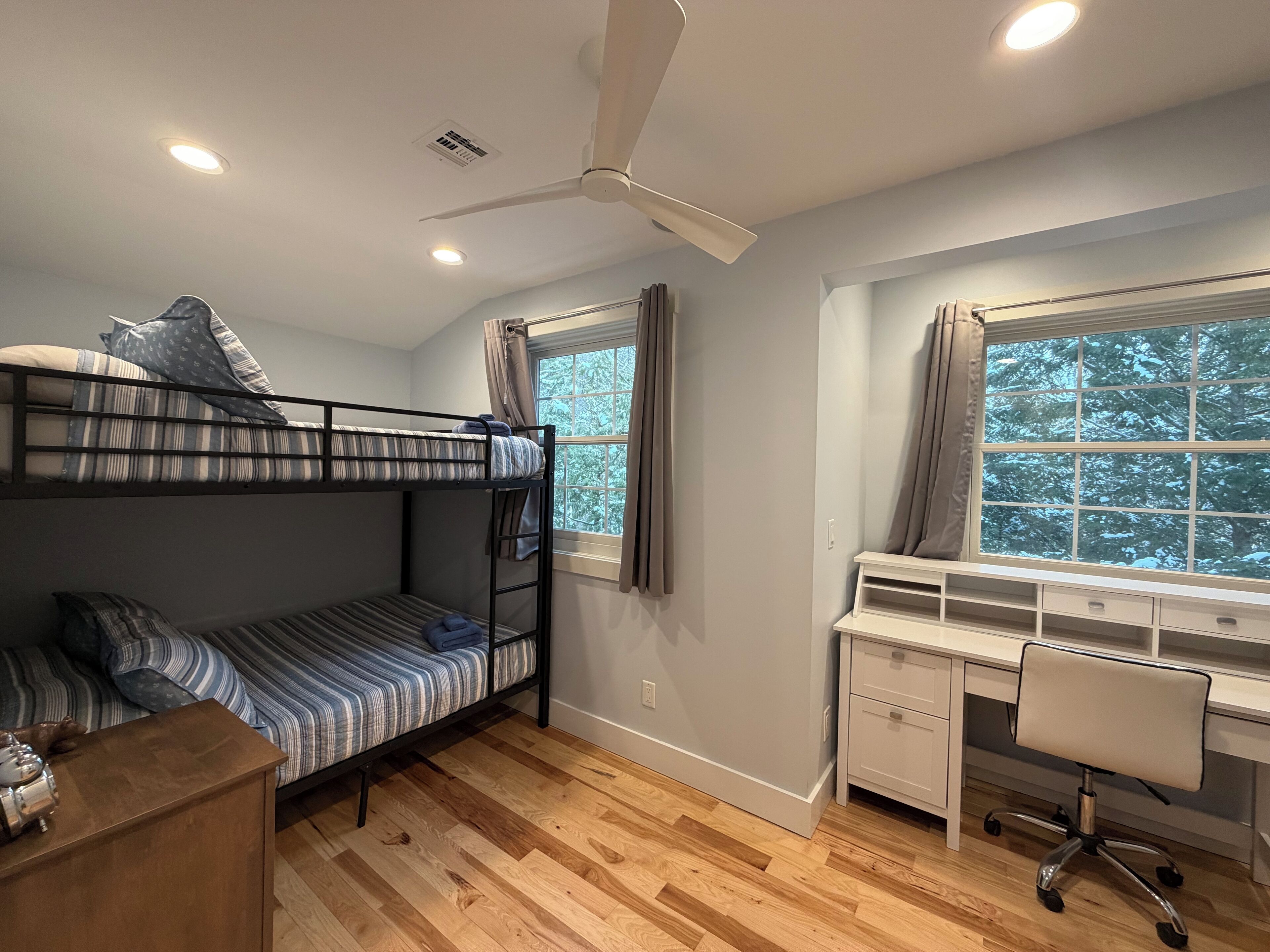 Un lit superposé avec un bureau et une chaise, un ventilateur de plafond et une fenêtre avec des rideaux.