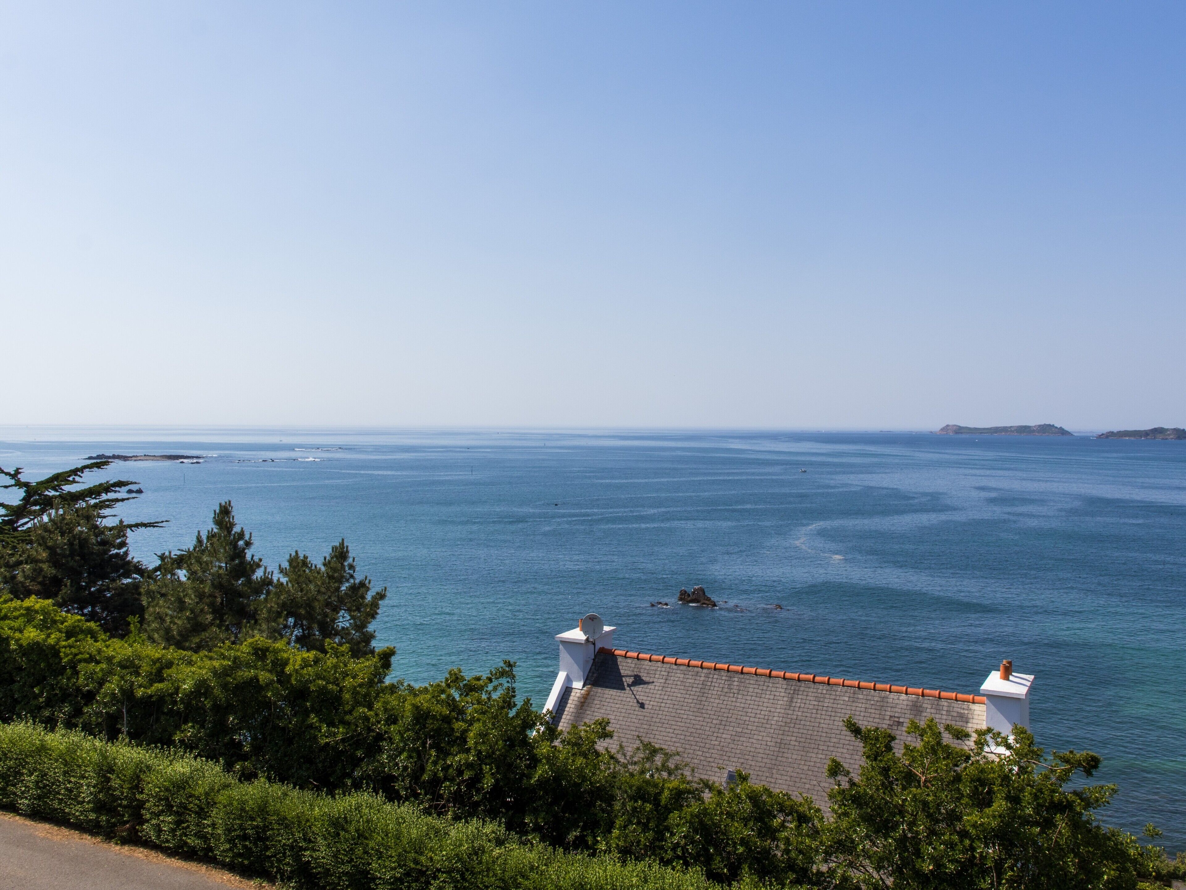 A coastal view with a house featuring a red-tiled roof, surrounded by greenery and overlooking the sea.