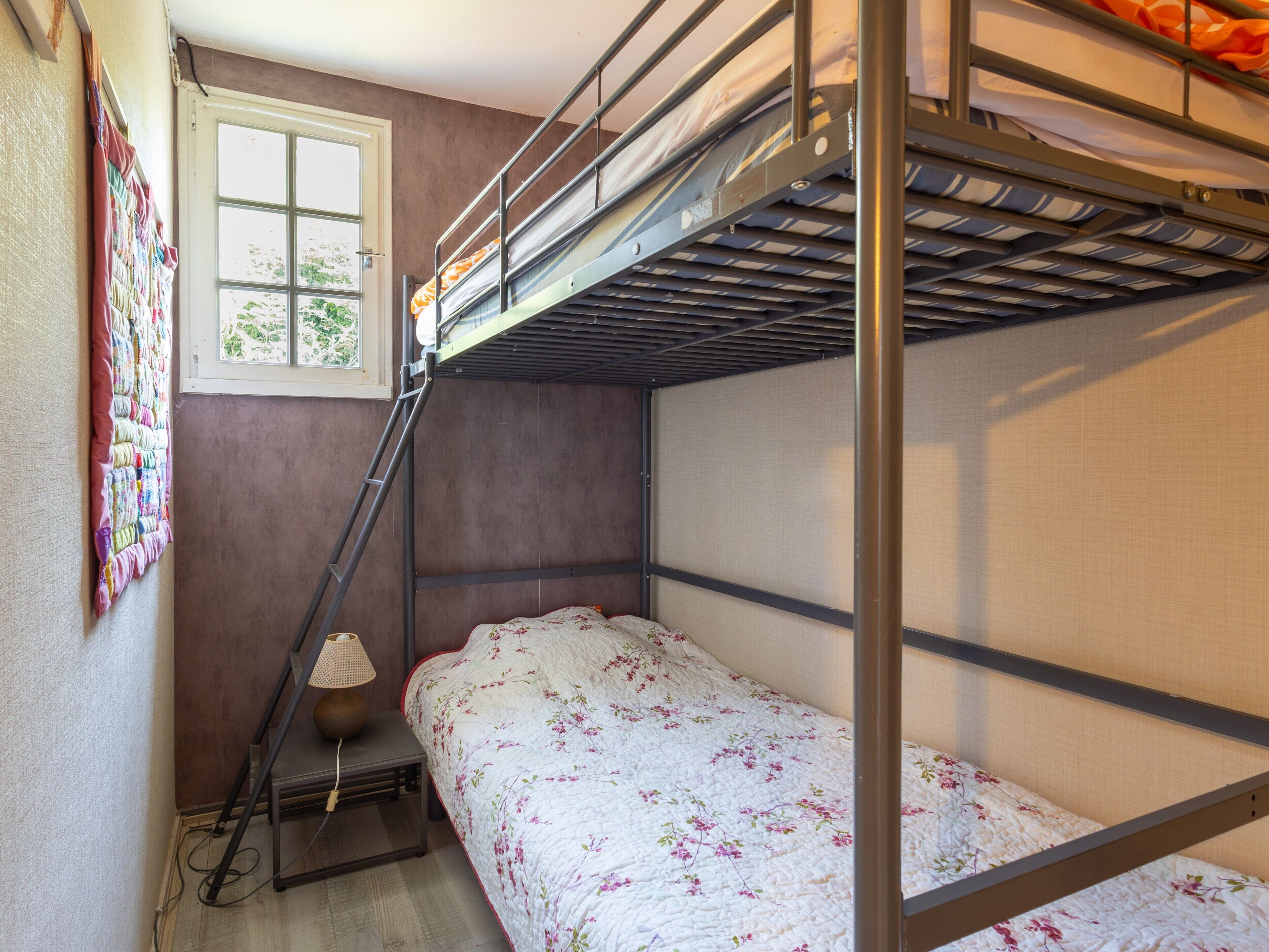 Une chambre avec un lit superposé, une fenêtre, une lampe et une courtepointe accrochée au mur.