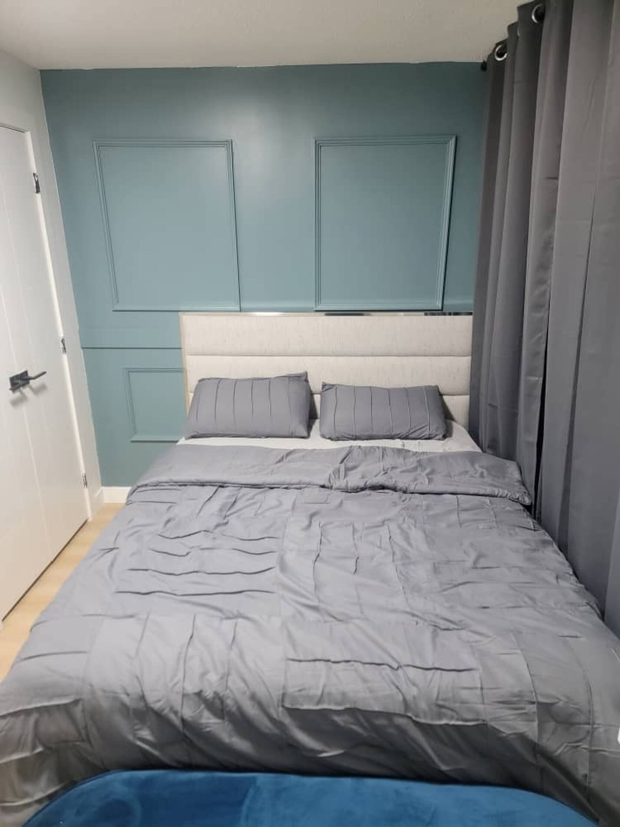 Une chambre avec une tête de lit de couleur turquoise, une literie grise et une porte située à gauche.