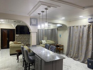 A modern kitchen with a marble countertop, bar stools, pendant lights, and a dining area with a sofa and chairs.