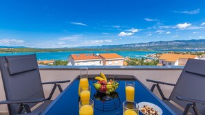 Un balcon avec une table préparée pour le petit-déjeuner, offrant une vue sur la côte, avec des bâtiments et des montagnes en arrière-plan.