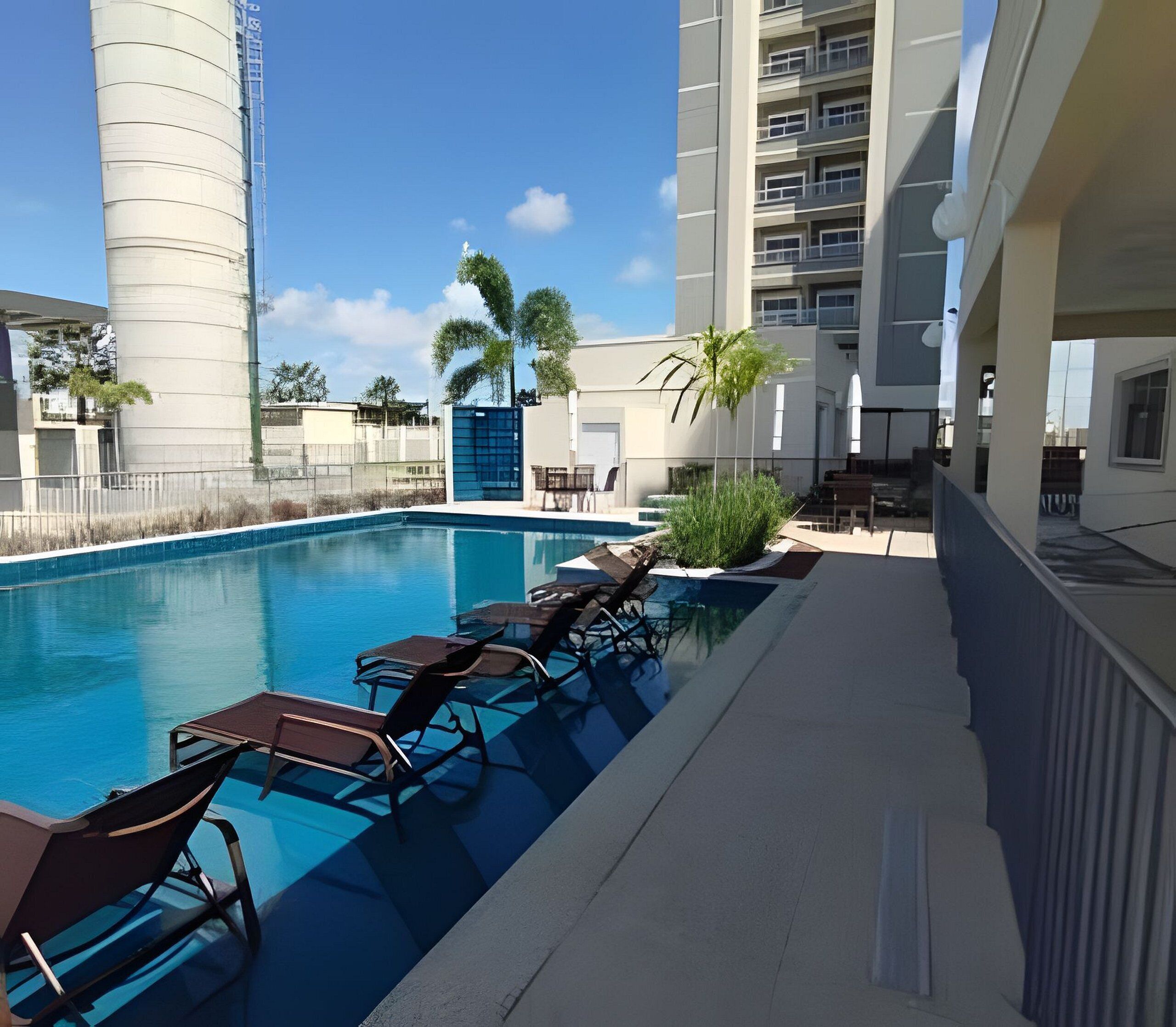Une piscine avec des chaises longues, un palmier et un grand immeuble en arrière-plan.