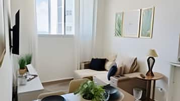 Uma área de refeições com uma mesa posta para quatro pessoas, um sofá, uma pequena mesa lateral com abajur e uma janela com cortinas.