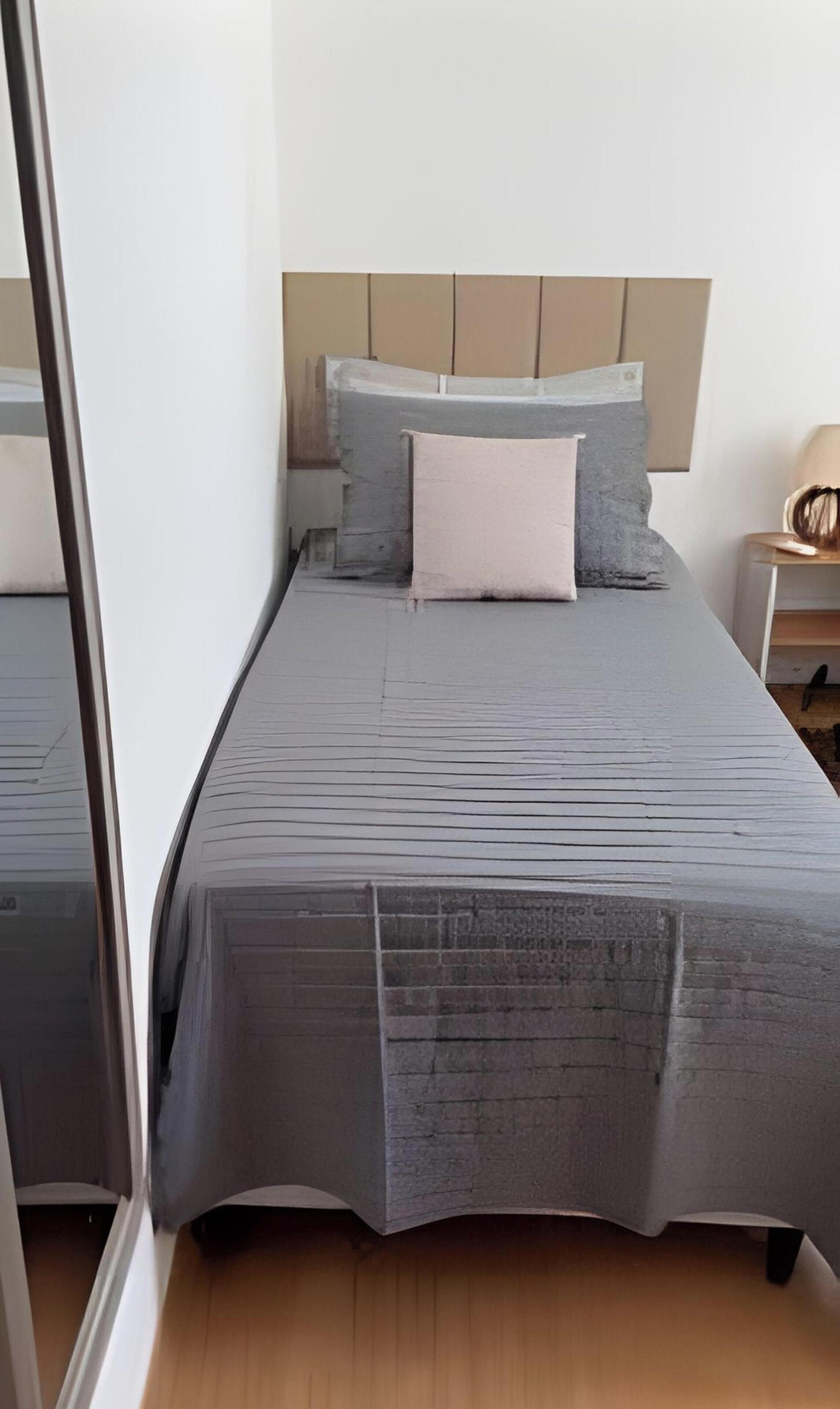 A single bed with a grey bedspread and two pillows, a wooden headboard, and a bedside table with a lamp.