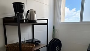 Una cucina con una scaffalatura metallica, una macchina da caffè, un bollitore, un cesto, uno sgabello e una finestra vista cielo.