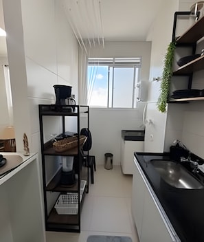 A compact kitchen with a black countertop, white cabinets, a small window, and a plant on the shelf. - 1505 Lazer conforto e tranquilidade (Cabedelo)