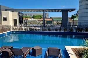 Una piscina con sillas de descanso, una zona cubierta y edificios al fondo.