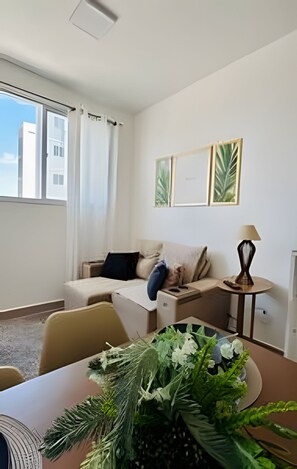 Una habitación con sofá, una mesita de noche con lámpara y una ventana con cortinas.