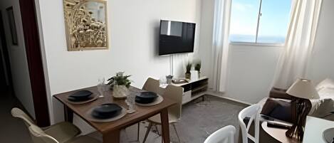 A modern living room with a dining area, a flat-screen TV mounted on the wall, and a large window with curtains.