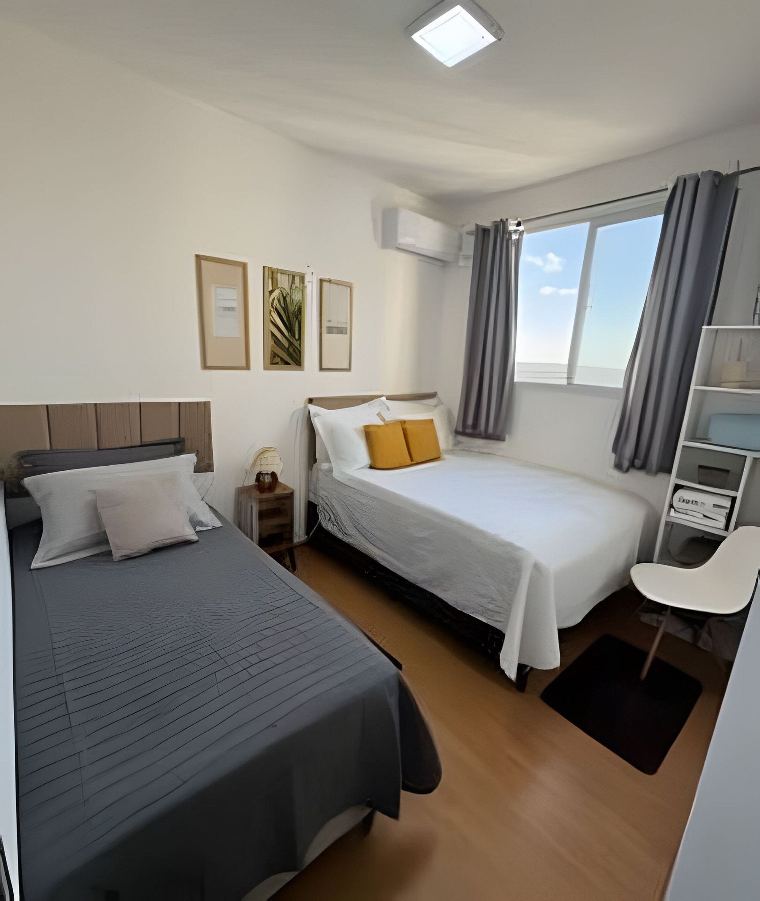 A hotel room with two beds, a desk, a chair, a window with curtains, and an air conditioning unit.