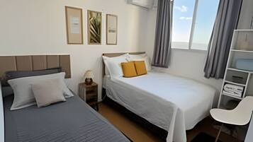 A hotel room with two beds, a desk, a chair, a window with curtains, and an air conditioning unit.