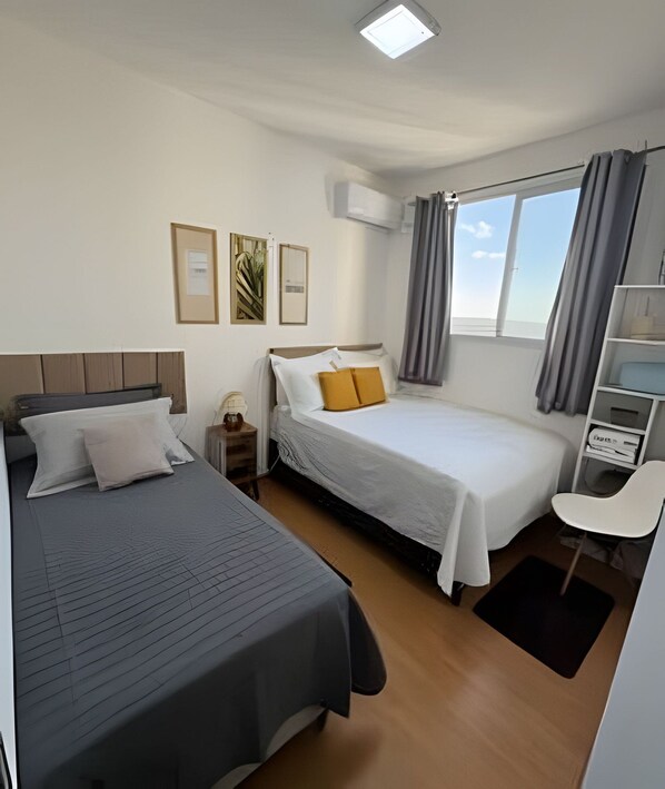 A hotel room with two beds, a desk, a chair, a window with curtains, and an air conditioning unit. - 1505 Lazer conforto e tranquilidade (Cabedelo)