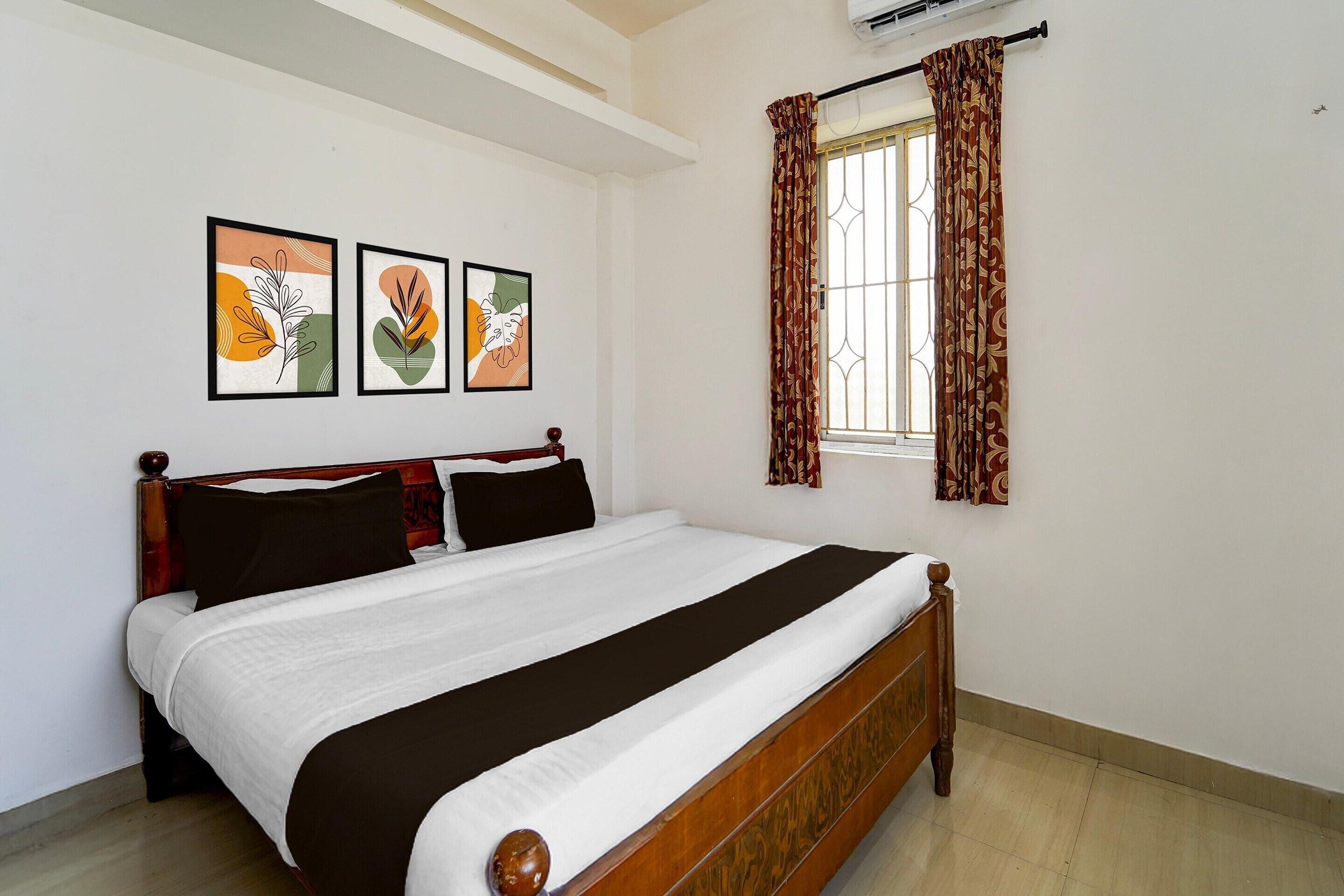 A neatly made bed with a wooden frame, a window with patterned curtains, and two framed floral artworks on the wall.