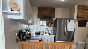 Cocina compacta con muebles blancos, una isla central, dos sillas de madera y un refrigerador de acero inoxidable.