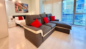 A modern living room with a sectional sofa, a small white bed, and a large window offering a city view. - Oshun Estudio Deluxe D312 (Madrid)
