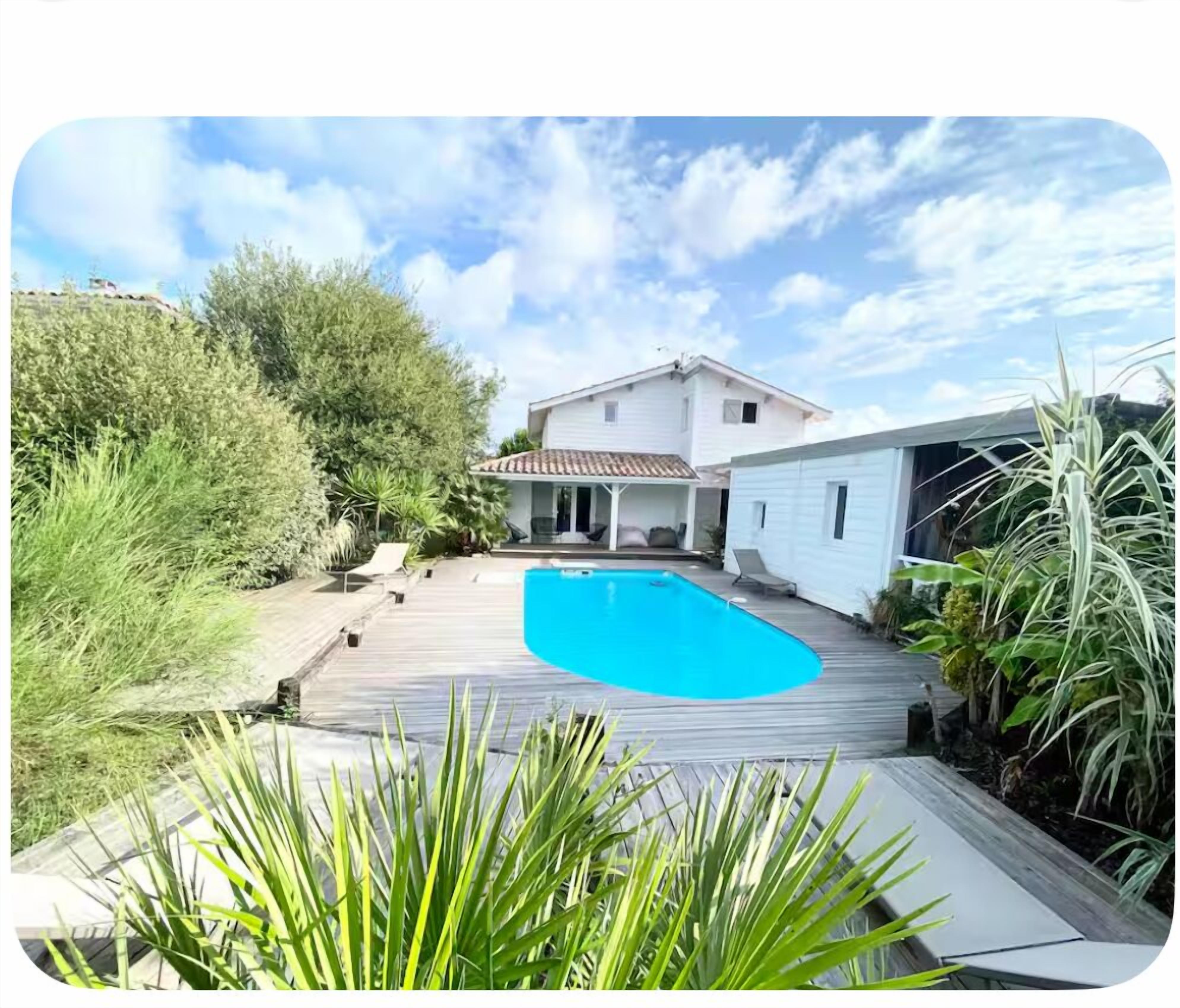 A pool area with a wooden deck, a house with a tiled roof, and surrounding greenery.