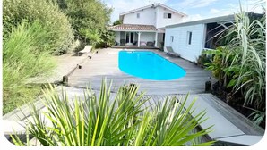 A pool area with a wooden deck, a house with a tiled roof, and surrounding greenery. - Private air-conditioned villa with heated pool 2nd line Bassin d'Arcachon (Gujan-Mestras)