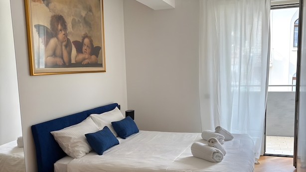 Une chambre avec un grand lit, un petit tabouret, un tableau accroché au mur et une vue par la fenêtre.