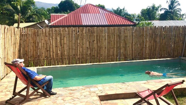 Ein Swimmingpool mit Bambuszaun, ein Gebäude mit rotem Dach und zwei Liegestühle.
