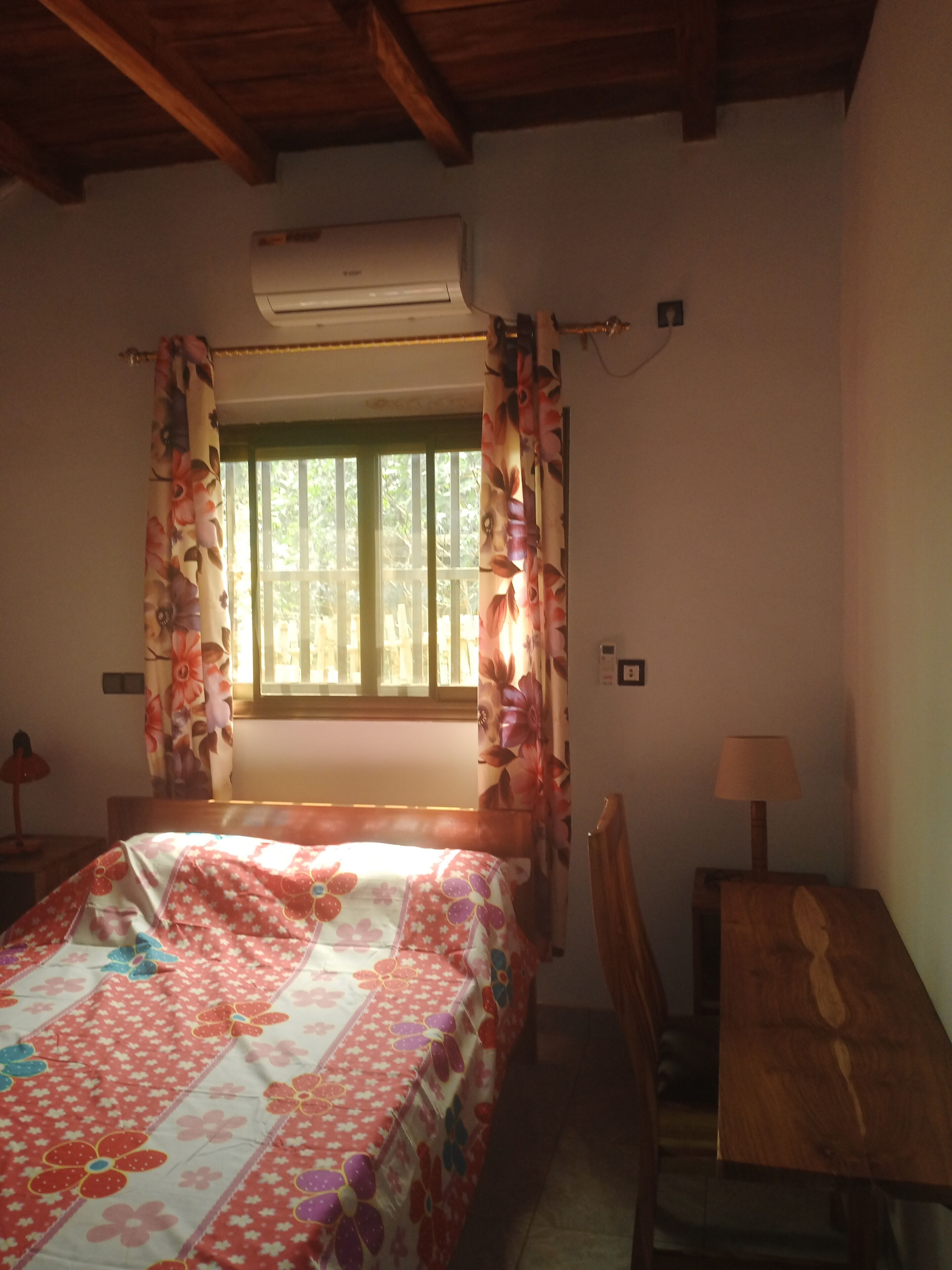 Een kamer met een bed, een raam met gordijnen, een airconditioningsunit, een bureau met lamp en een stoel.